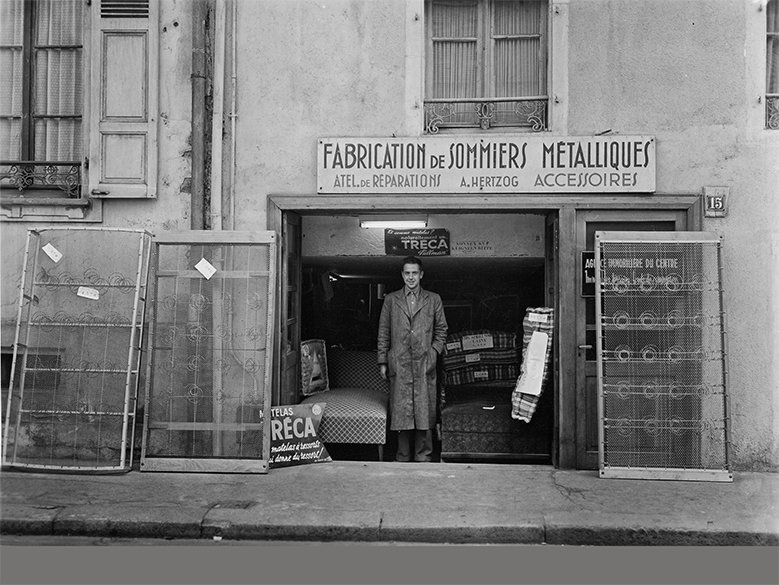 Ancienne photo en noir et blanc du premier point de vente d'Hertzog avec un homme devant et des sommiers de la marque Treca